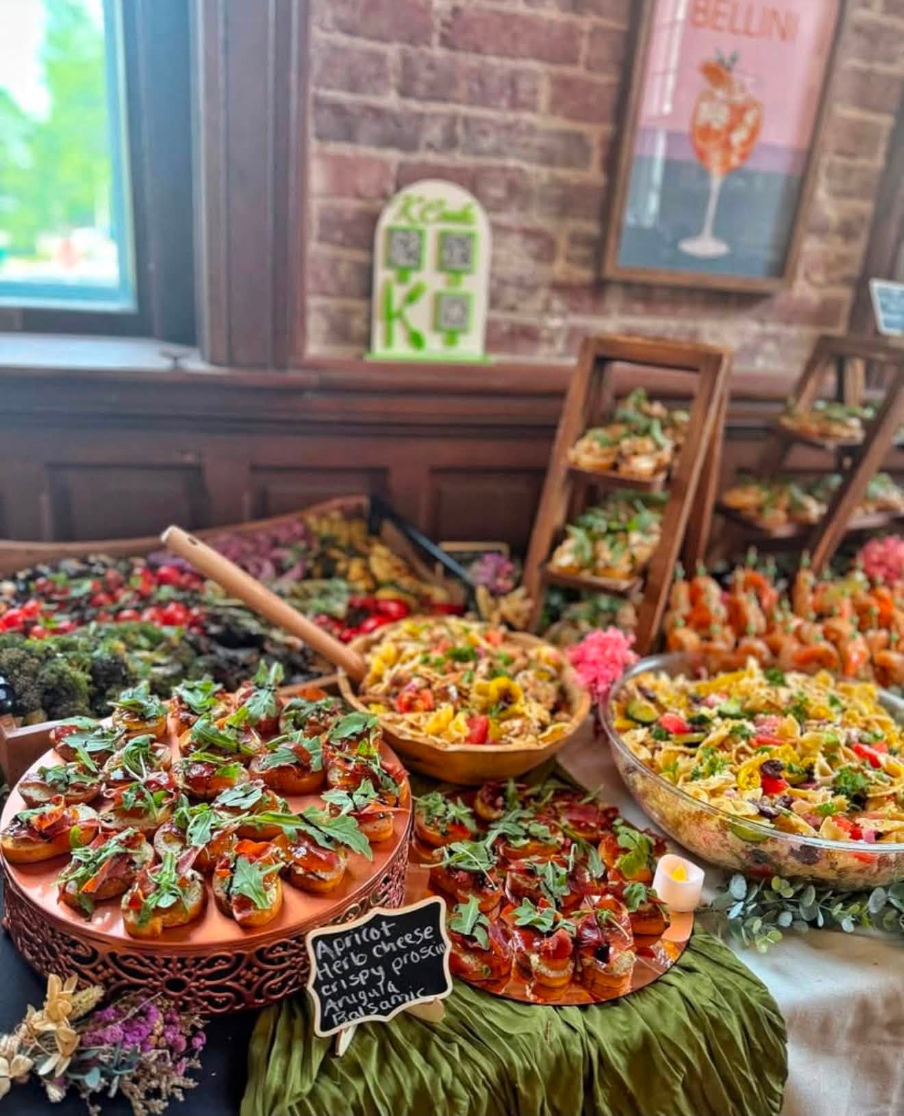 Large K Cooks event buffet with crostini, pasta salads, and shrimp in front of a brick wall