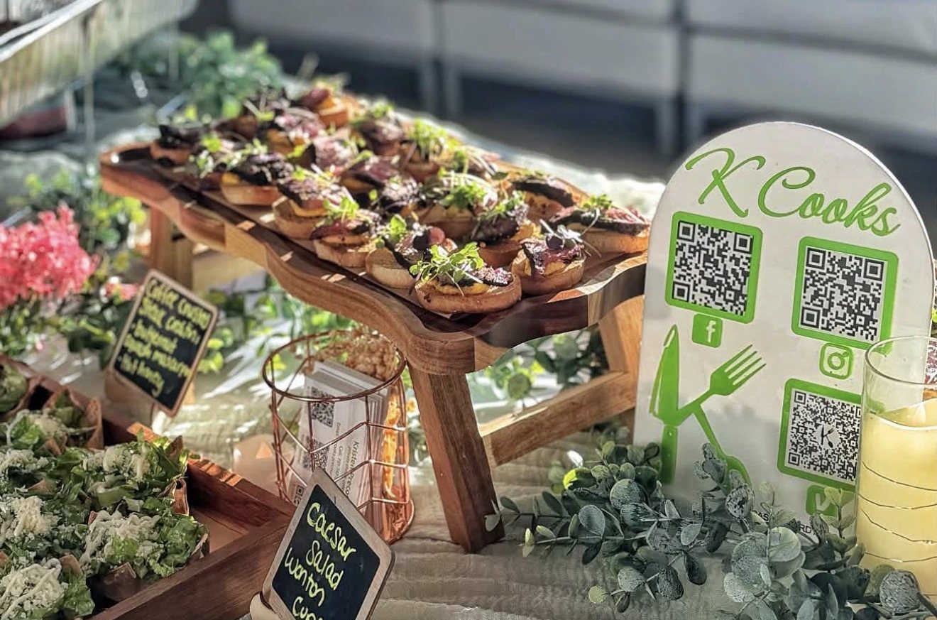 Styled catering display of croissant sandwiches on a wooden riser with a K Cooks QR sign and greenery
