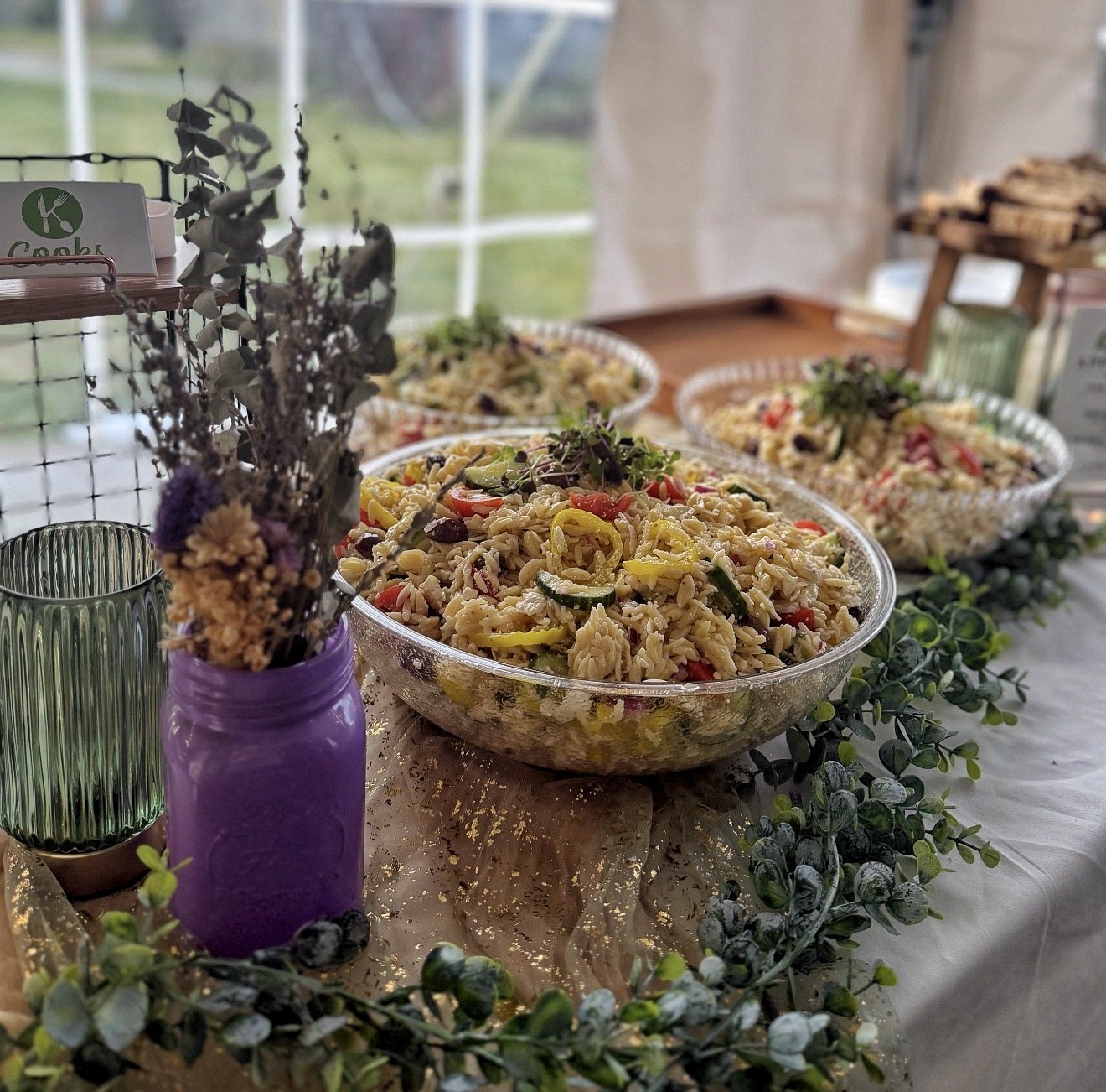 Large bowls of pasta salad on a display table with greenery and a jar of purple flowers