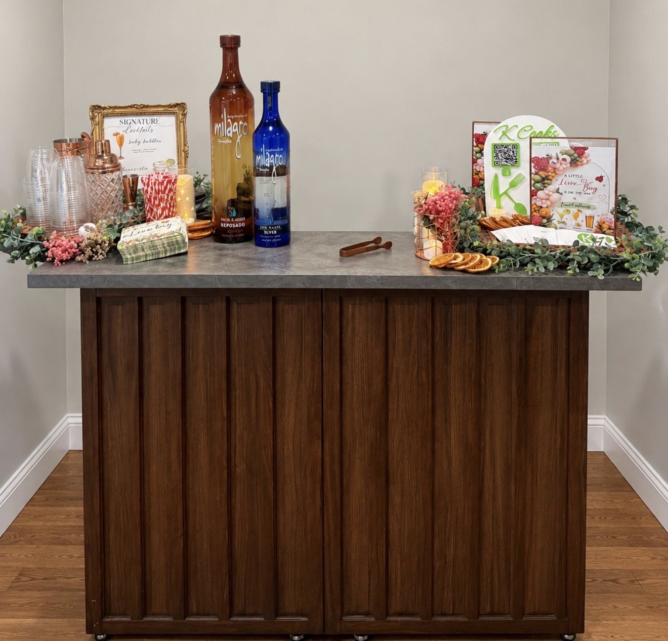 K Cooks bar setup with liquor bottles, glassware, cocktail tools, greenery, and a K Cooks sign